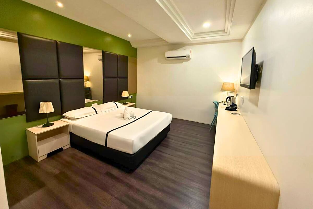 A modern hotel room with a large bed, a bedside table, a nightstand, a mirror, a flat-screen TV mounted on the wall, and an air conditioning unit.