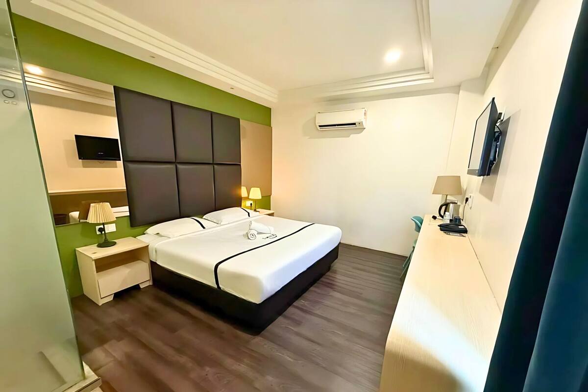 A hotel room with a bed, bedside tables, a TV, an air conditioning unit, and a desk with a lamp.