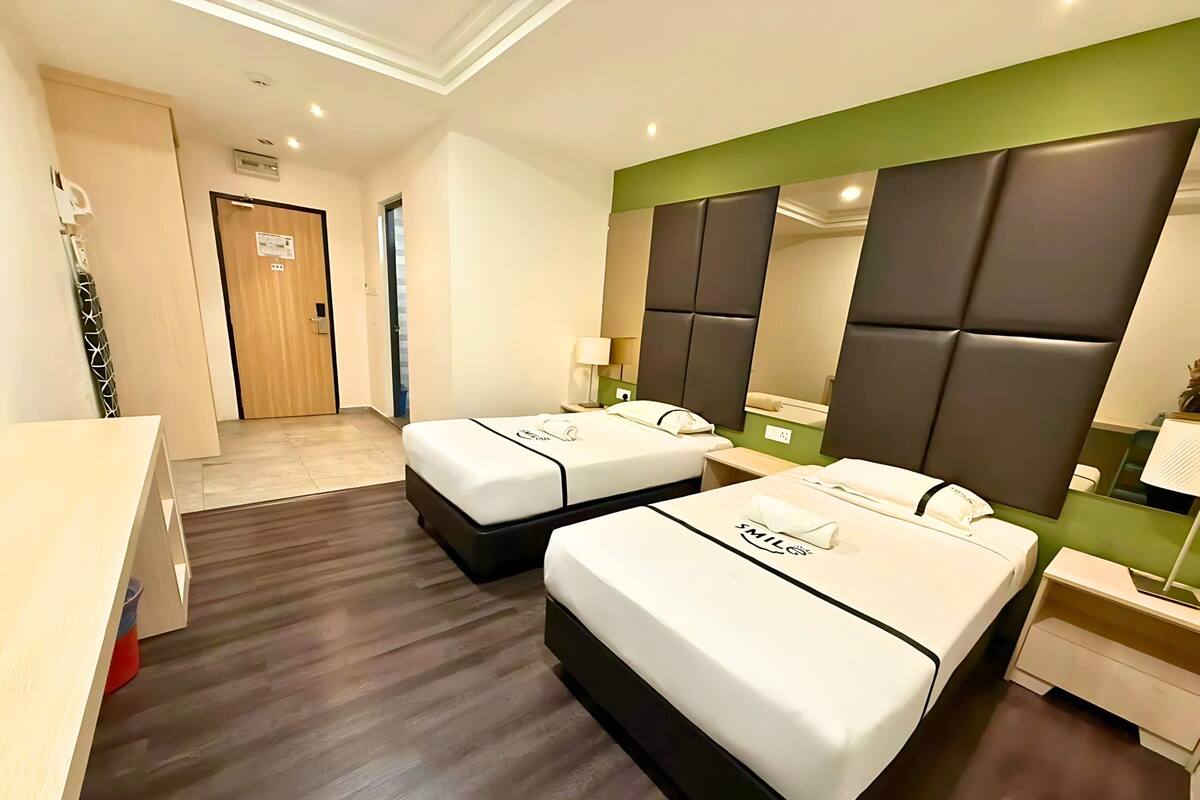 A hotel room with two beds, a desk, a mirror, and a green accent wall.