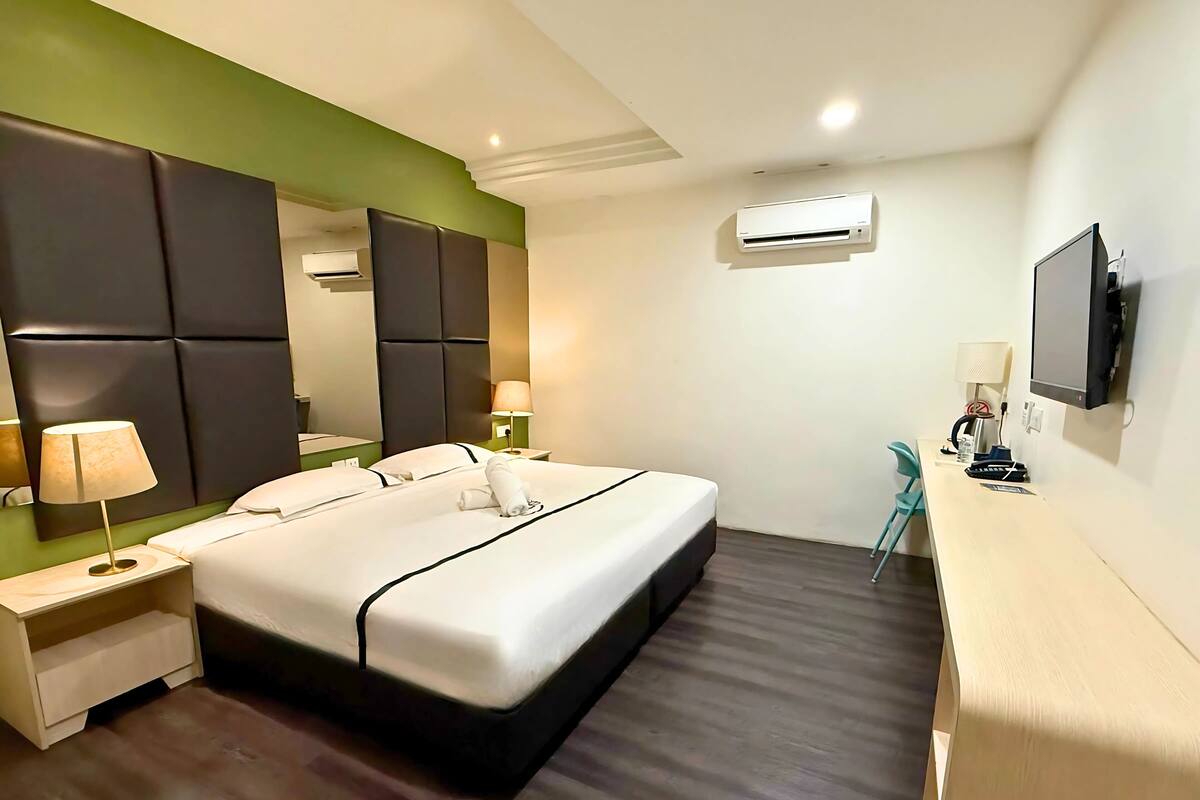A hotel room with a large bed, a desk, a chair, a TV, and an air conditioning unit.