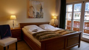 A hotel room with a bed, a chair, a nightstand, a lamp, a poster, and a window with a view of buildings. - Bodenschneid Suites Rottach-Egern - Bodenschneid Suites Wallbergblick (Rottach-Egern)