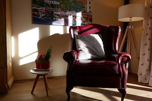 Un sillón rojo de cuero con un cojín estampado, una mesita con una planta, una lámpara y una pared con un cartel de colores.