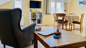 Un salon confortable avec un canapé, une table à manger et des chaises, un téléviseur fixé au mur et une bibliothèque.