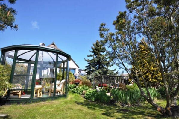 A glass greenhouse structure with a roof, surrounded by a garden with various plants and a tree.