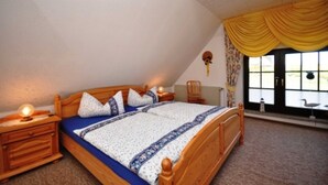 Ein Schlafzimmer mit einem hölzernen Bett, einem Nachttisch, einem Fenster mit gelben Vorhängen und Blick ins Freie.