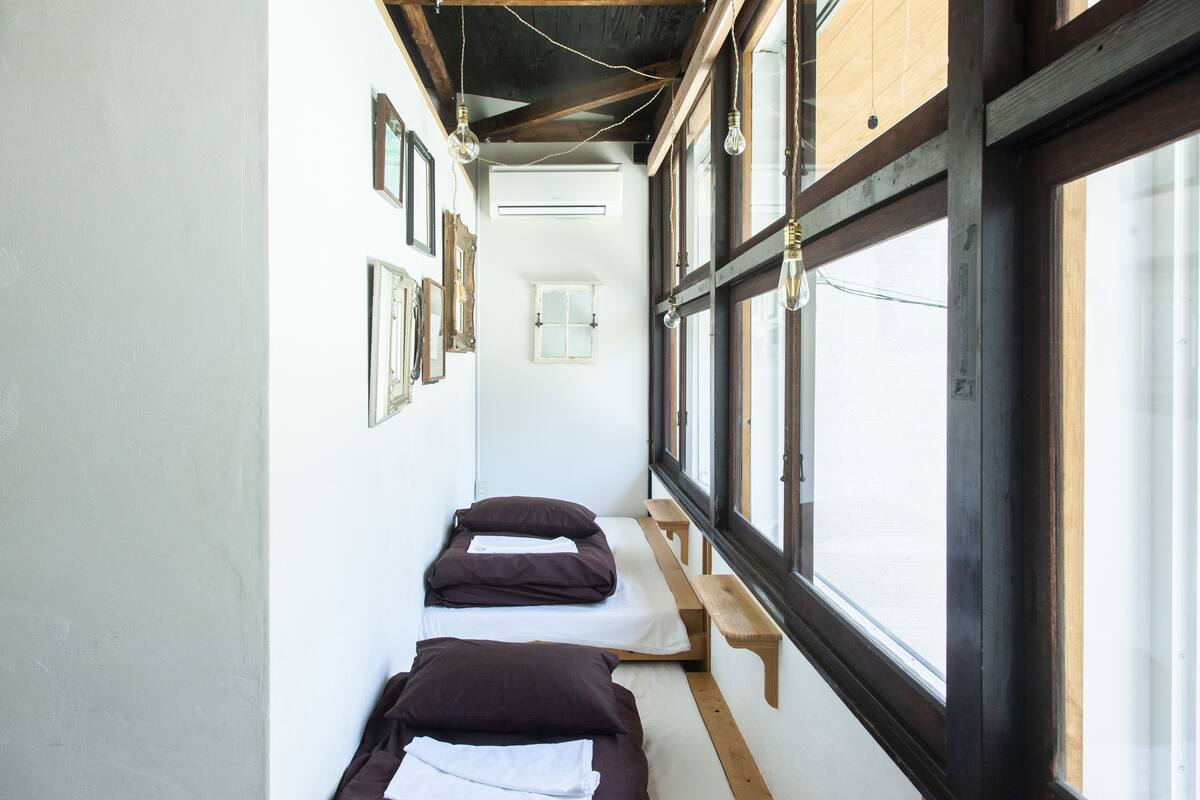 Un couloir étroit avec deux lits alignés contre le mur, des bancs en bois et de grandes fenêtres laissant entrer la lumière naturelle.