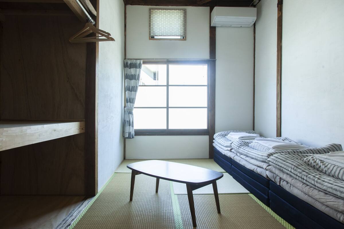 Une pièce avec un sol recouvert de tatamis, un futon servant de lit, une petite table, une fenêtre avec un rideau et une structure en poutres de bois.