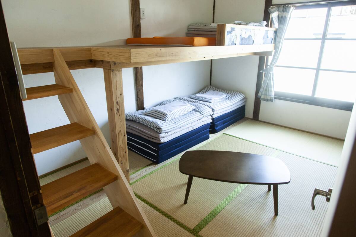 Une pièce avec un lit superposé, une petite table, des tatamis et une fenêtre avec des rideaux.