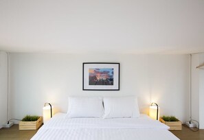 A minimalist bedroom with a large bed, two bedside tables with lamps, a framed picture on the wall, and two wooden crates with plants. - Sleeping Well Chiangkhong - Hostel (Chiang Khong)