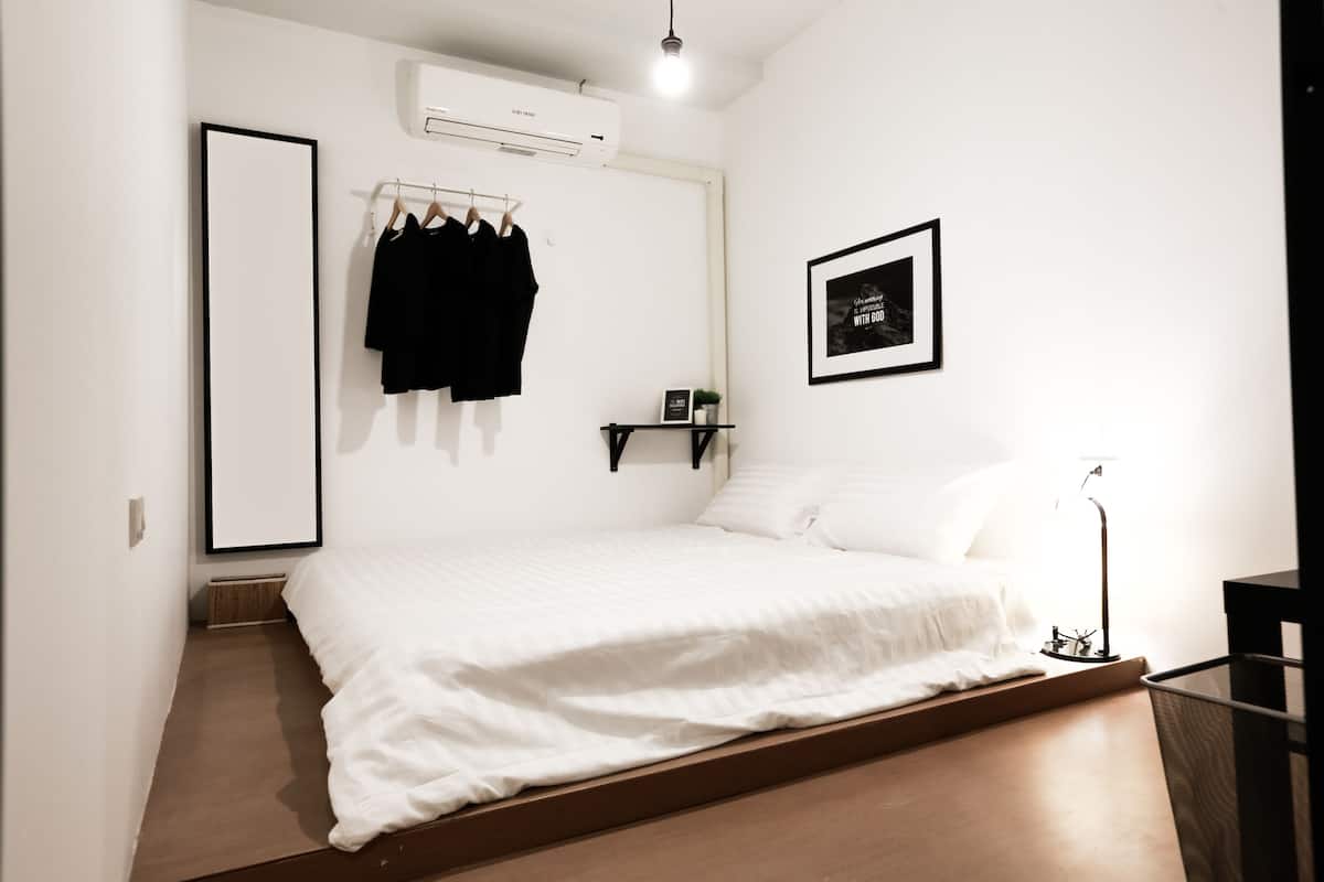 Ein schlicht eingerichtetes Schlafzimmer mit Bett, Wandklimaanlage, schwarzem Kleiderhaken, gerahmtem Bild und Nachttisch mit Lampe.