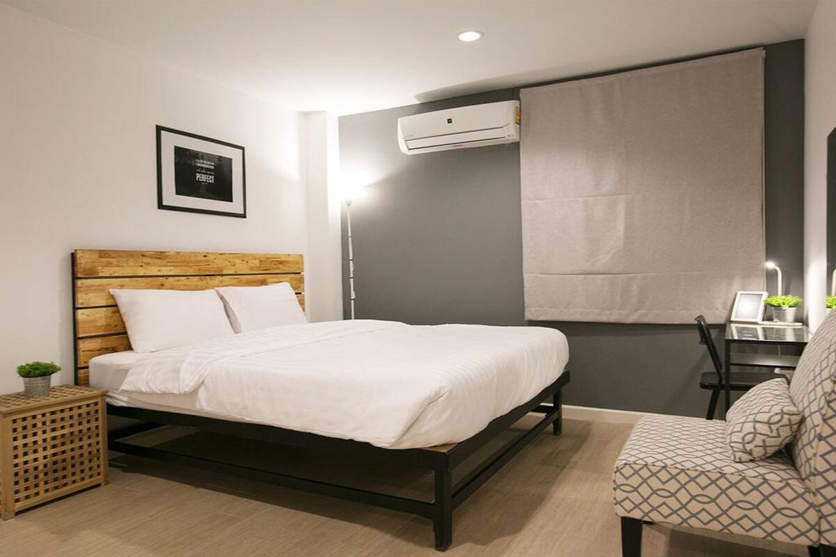 Ein modernes Schlafzimmer mit einem Bett mit Holz-Kopfteil, einem kleinen Nachttisch mit Pflanze, einer grauen Wand und einem Fenster mit weißem Rollo.