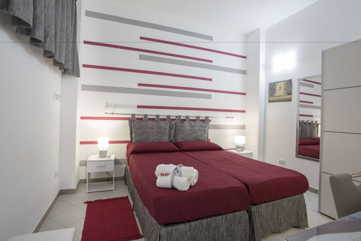 Une chambre d’hôtel moderne avec un lit soigneusement fait, une table de chevet avec une lampe, un tapis rouge et un grand miroir.