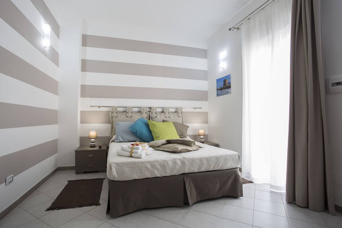 Une chambre à coucher bien rangée, avec un mur à rayures, un lit avec des oreillers, des lampes de chevet et une fenêtre avec des rideaux.