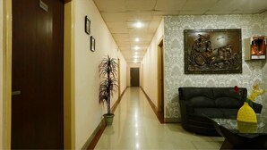 A hotel corridor with a wooden door, a potted plant, a framed picture, a sofa, and a decorative metal sculpture. - Hotel Green Lotus Dwarka (New Delhi)