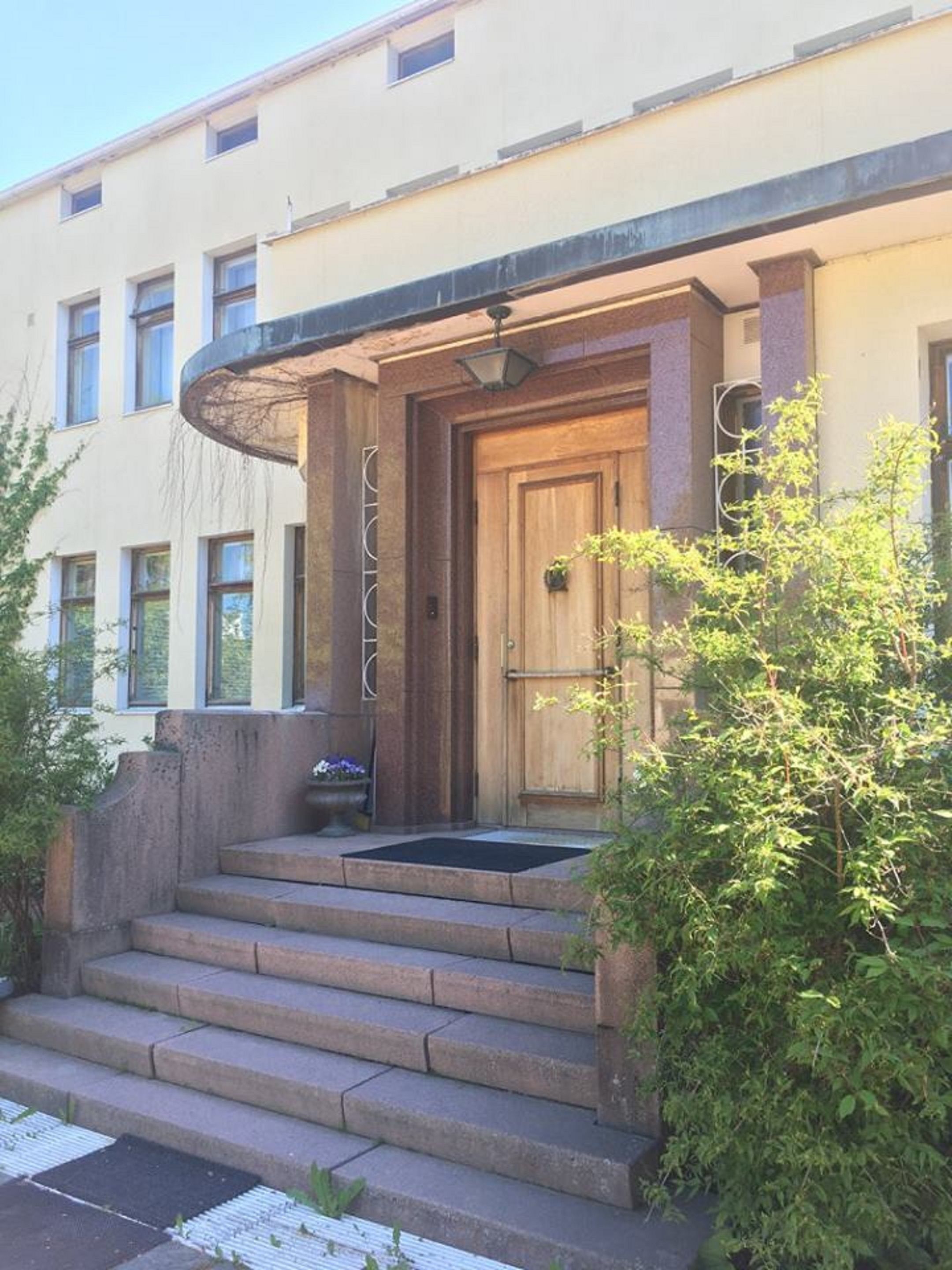 Rakennuksessa on puuovi, kivestä tehdyt portaat, jotka johtavat sisäänkäynnille, sekä pieni kukkaistutus.