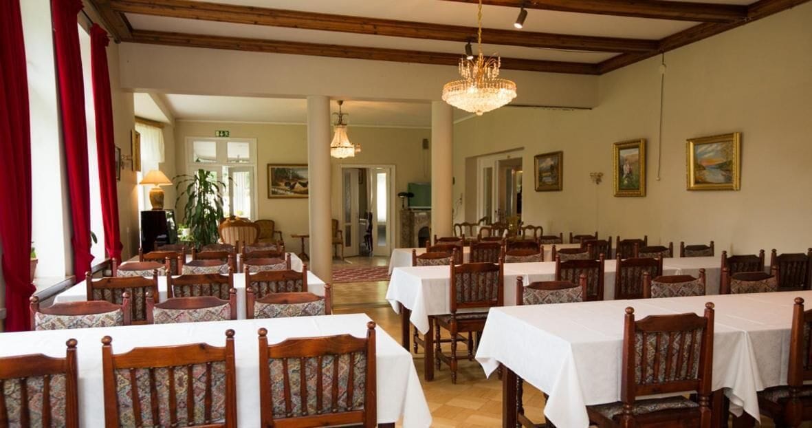 Een eethoek met meerdere tafels en stoelen, houten balken in het plafond en een kroonluchter.