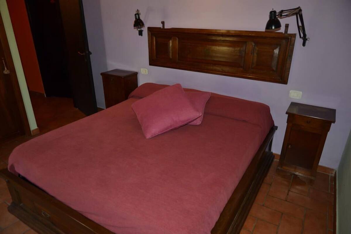 A bedroom with a wooden bed frame, a red bedspread, a single pink pillow, two bedside tables, and a nightstand.