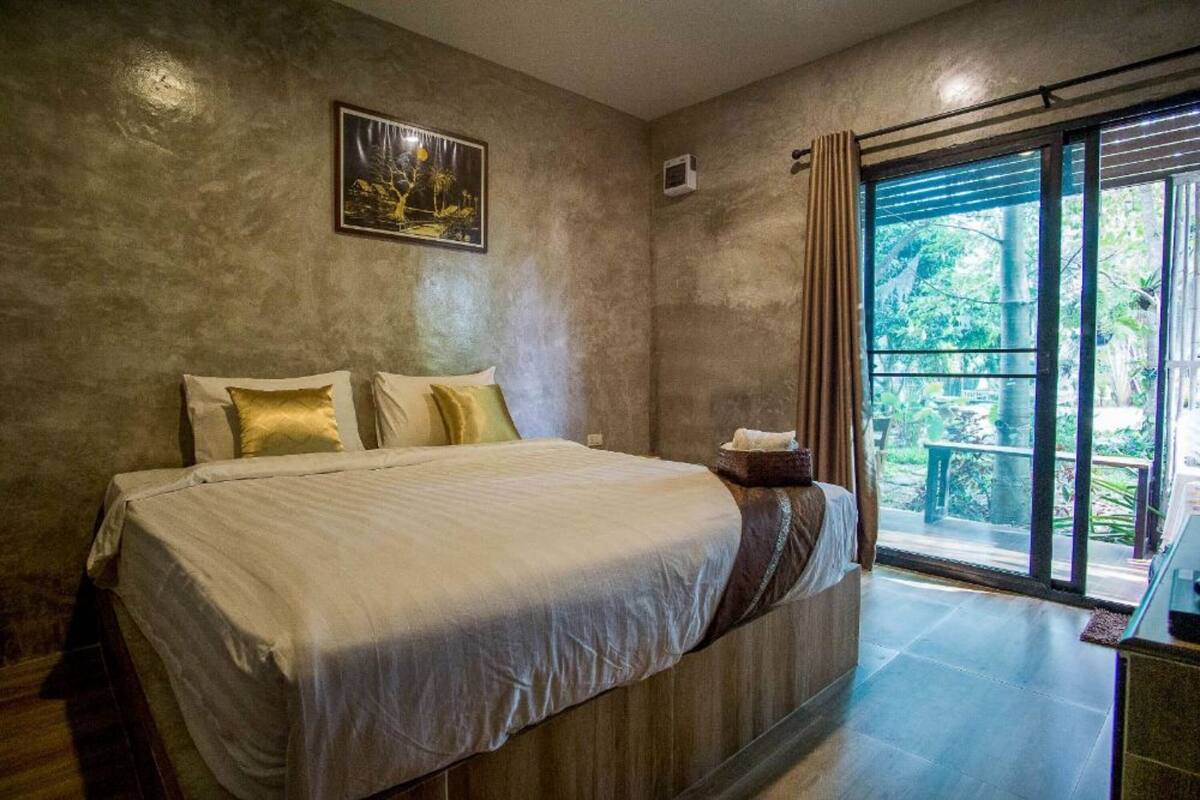 A bedroom with a large bed, a view of greenery through a window, and a painting on the wall.
