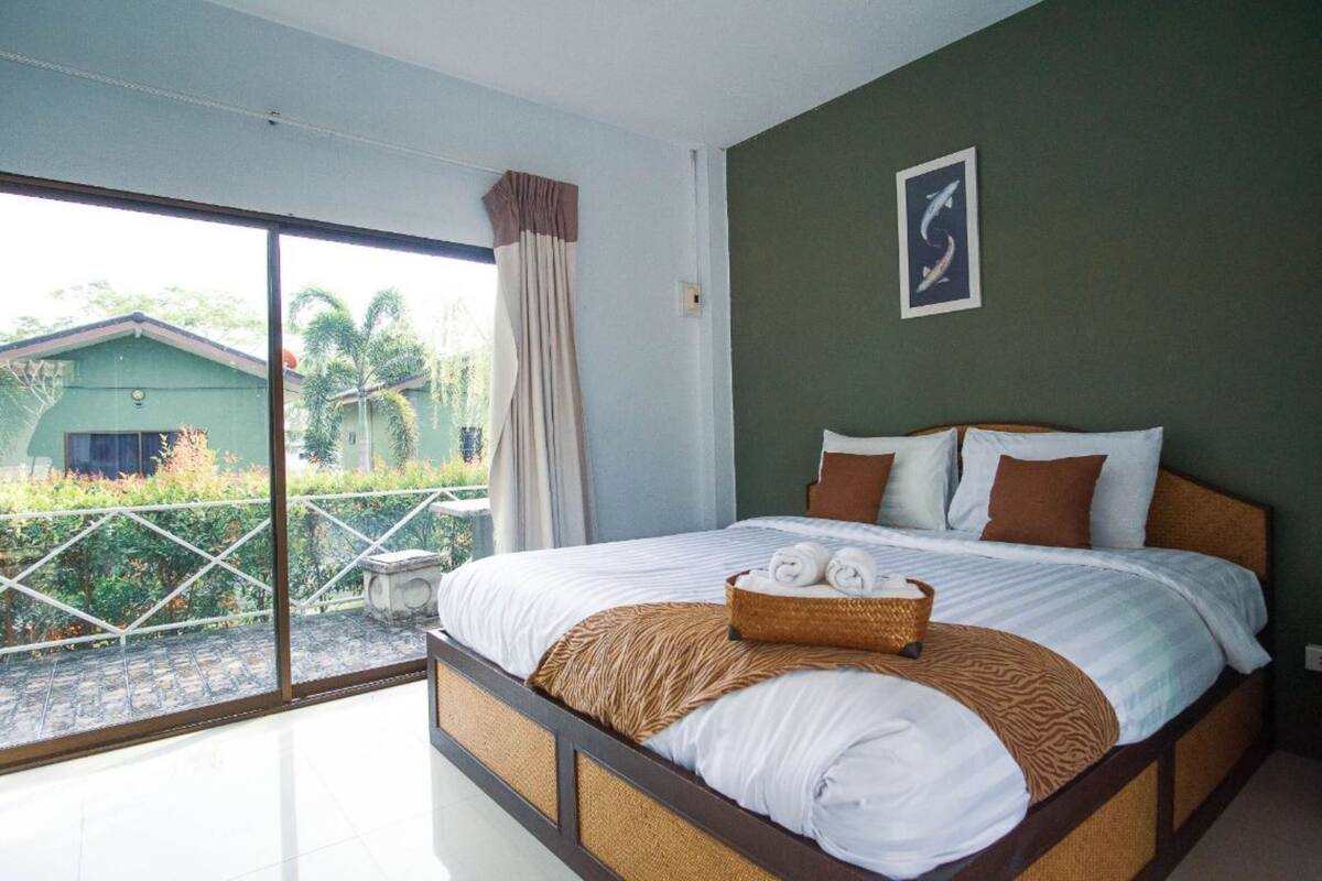 A bedroom with a large bed, a view of a green house, and a balcony with a railing.