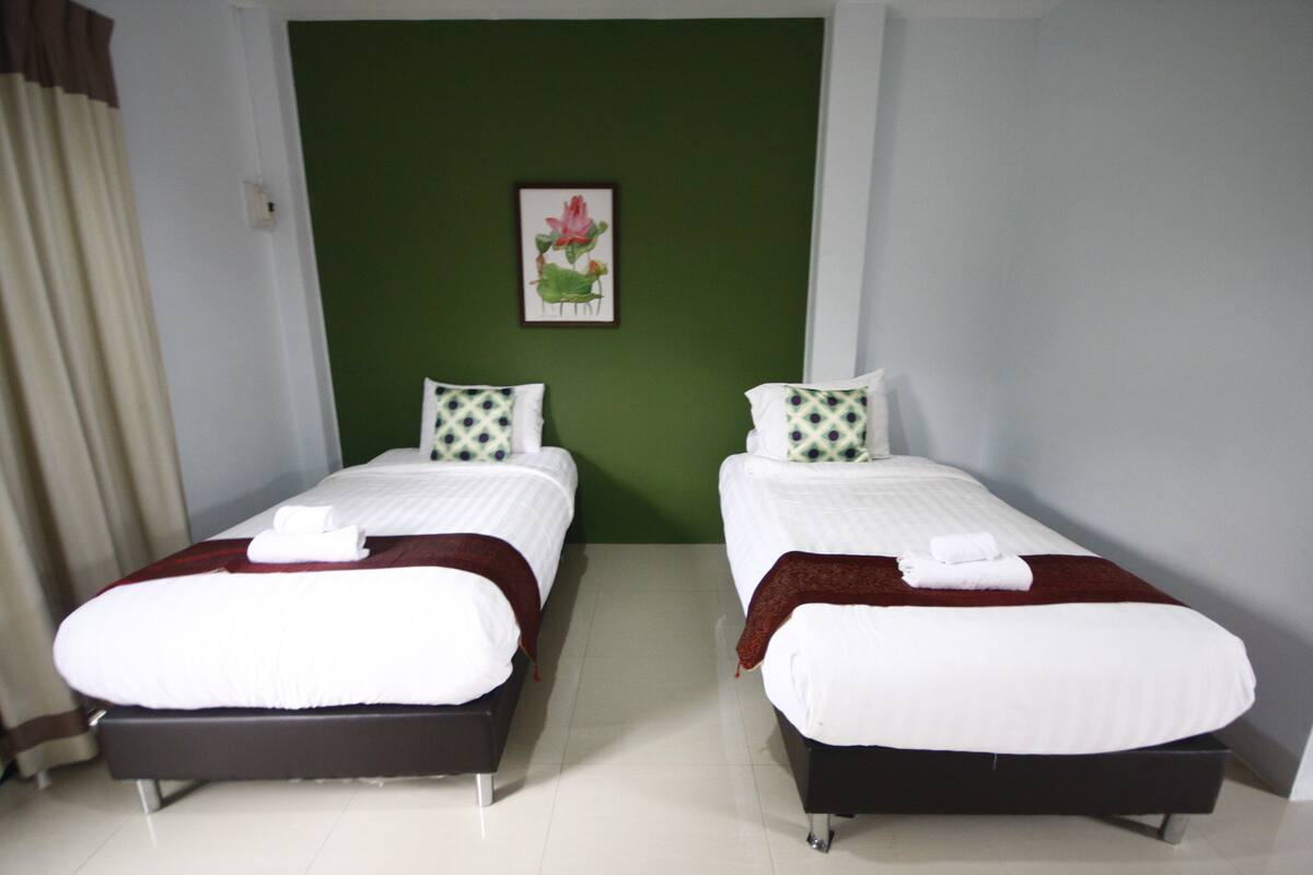A hotel room with two single beds, each with white bedding and a brown throw blanket. The room has a green accent wall and a framed picture of flowers.