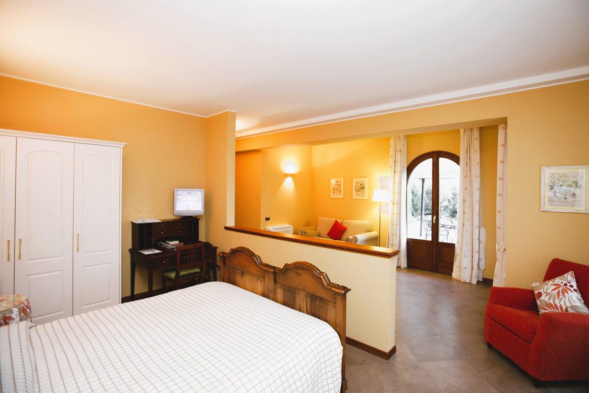 Habitación de hotel con cama, escritorio con computadora, sillón rojo y vista a través de un arco.