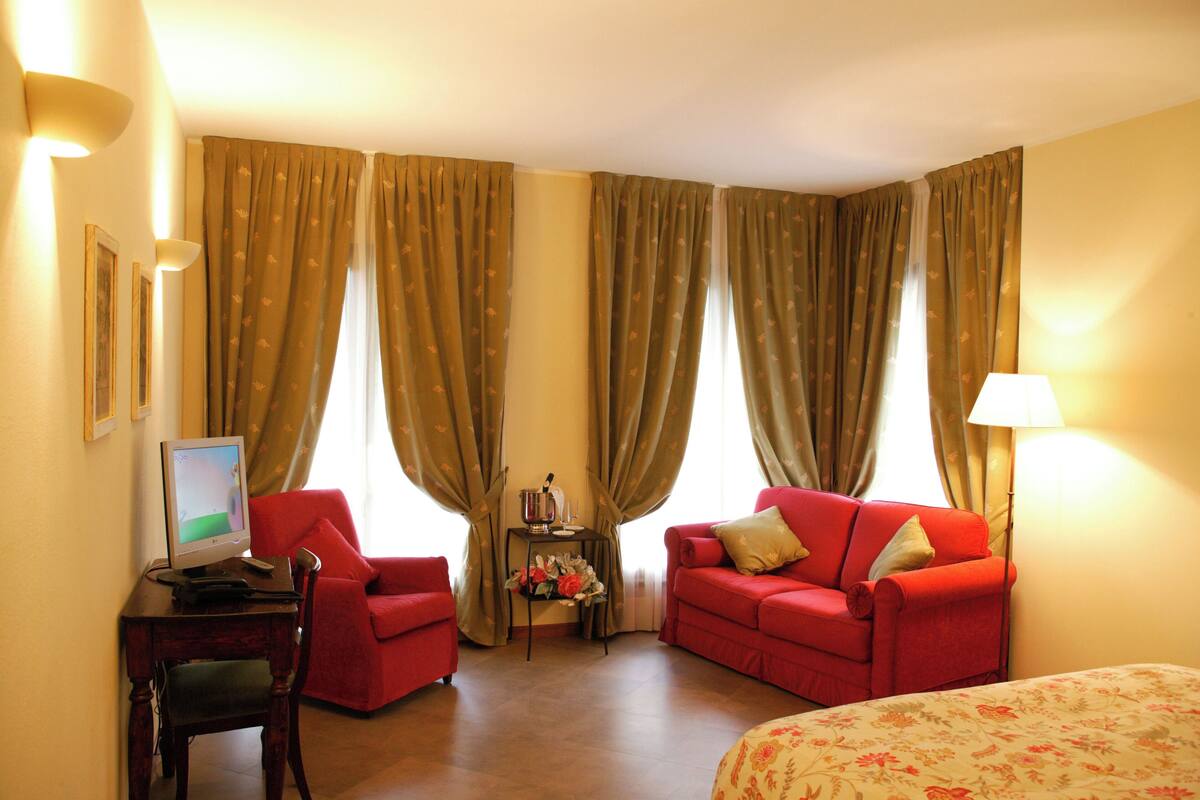 Habitación de hotel con un sofá rojo, una mesita con una lámpara, un televisor y cortinas.