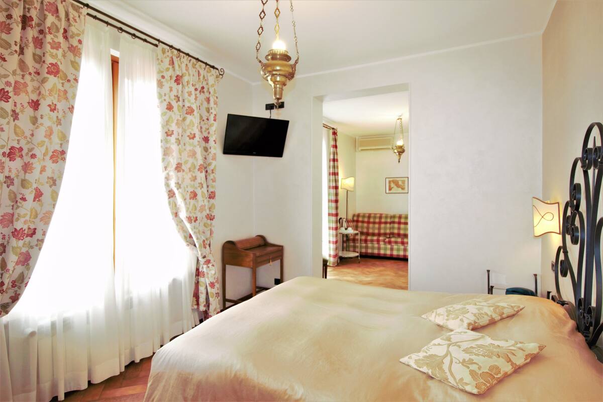Habitación de hotel con cama, televisión en la pared y vista a otra habitación con un sofá a cuadros.