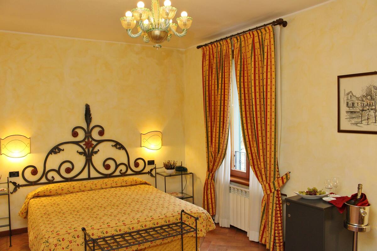 Un dormitorio con cama, candelabro, mesita de noche, radiador, ventana con cortinas y un cuadro enmarcado en la pared.