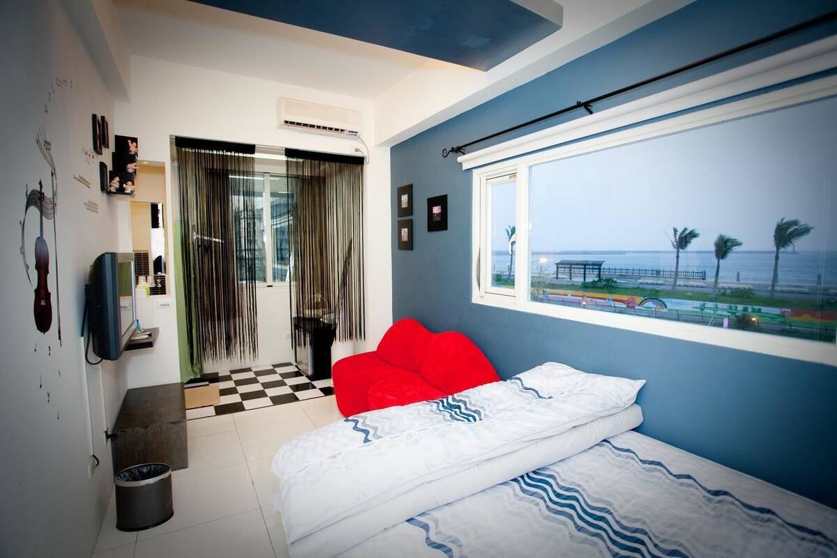 Un dormitorio con una cama, una silla roja, una ventana con vistas al mar y un suelo a cuadros.