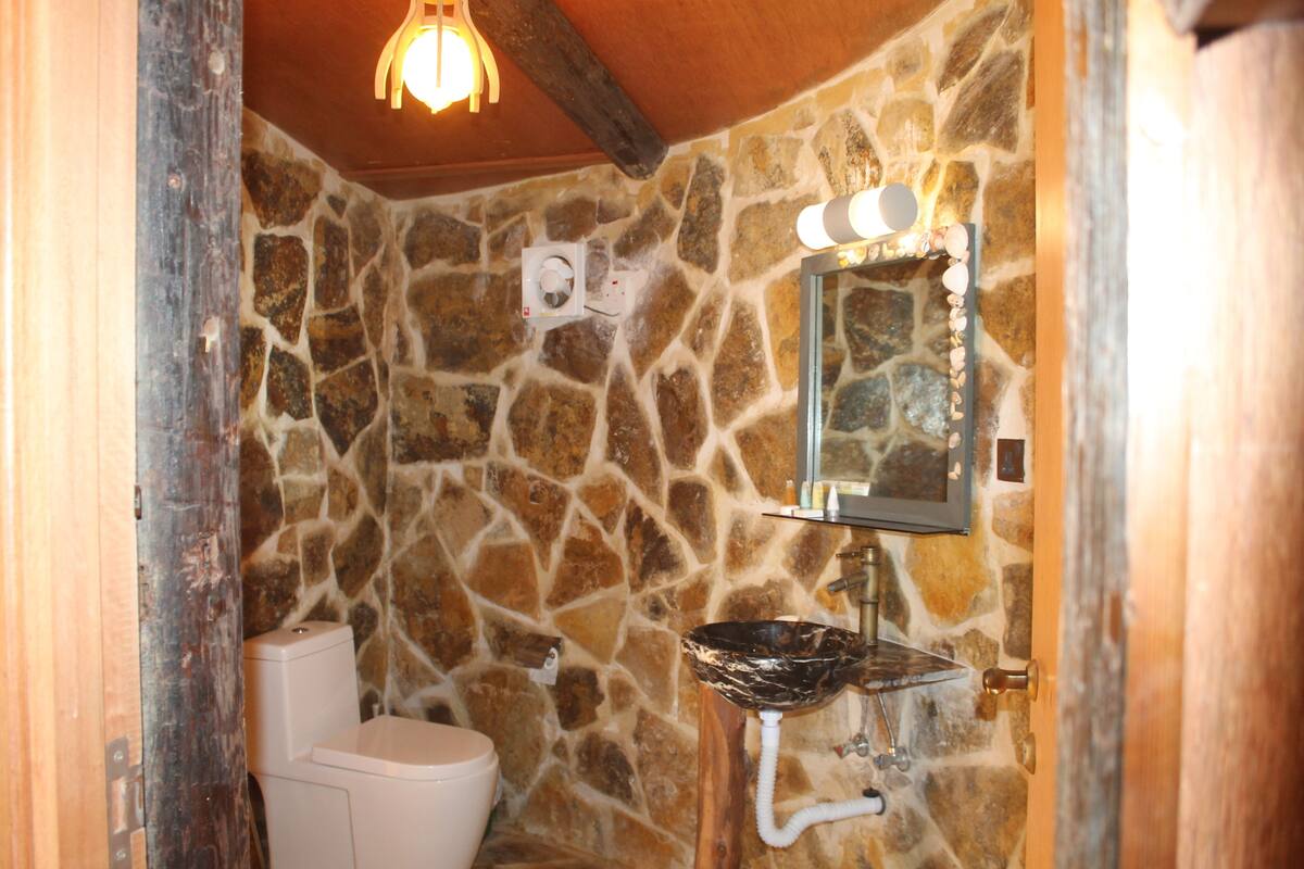A bathroom with a stone wall, a white toilet, a glass sink, and a mirror with lights.