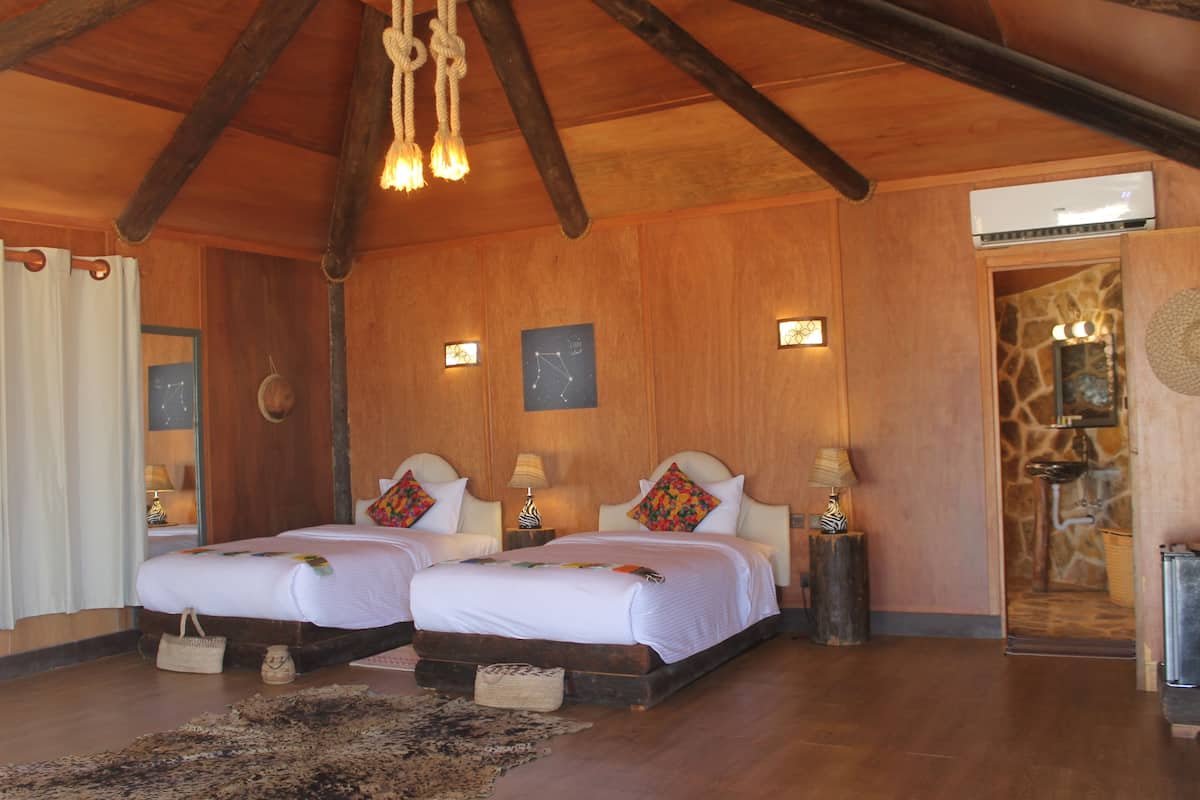 A spacious room with wooden beams, two beds with white linens, a stone wall, and a small bathroom visible through an open door.