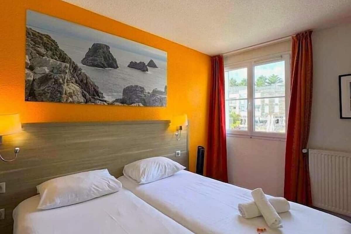 Ein Hotelzimmer mit zwei Betten, einem Gemälde einer felsigen Küste, einem Fenster mit Vorhängen und einem Heizkörper.