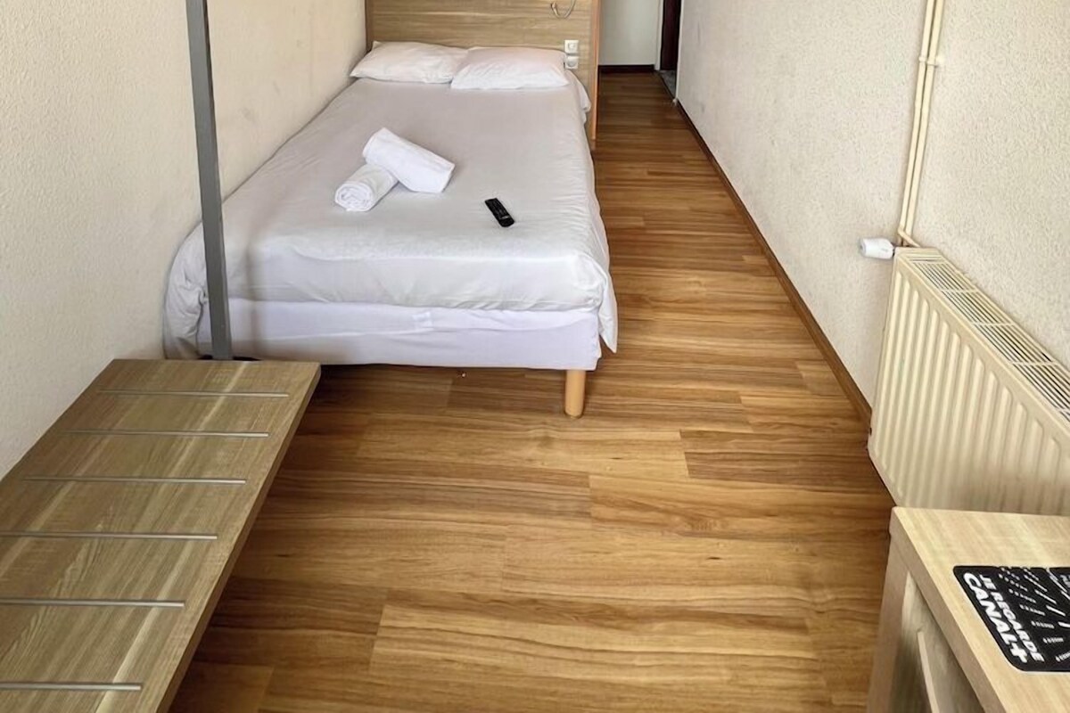 A narrow hotel room with a single bed, a wooden floor, a painting on the wall, and a small table with a lamp.