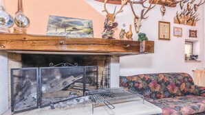 Ein Kamin mit Steinvorsatz, einem Metallgitter und einem hölzernen Regal darüber. Ein floral gemustertes Sofa und eine Wand mit verschiedenen Dekorationen, darunter Geweihe und gerahmte Urkunden.