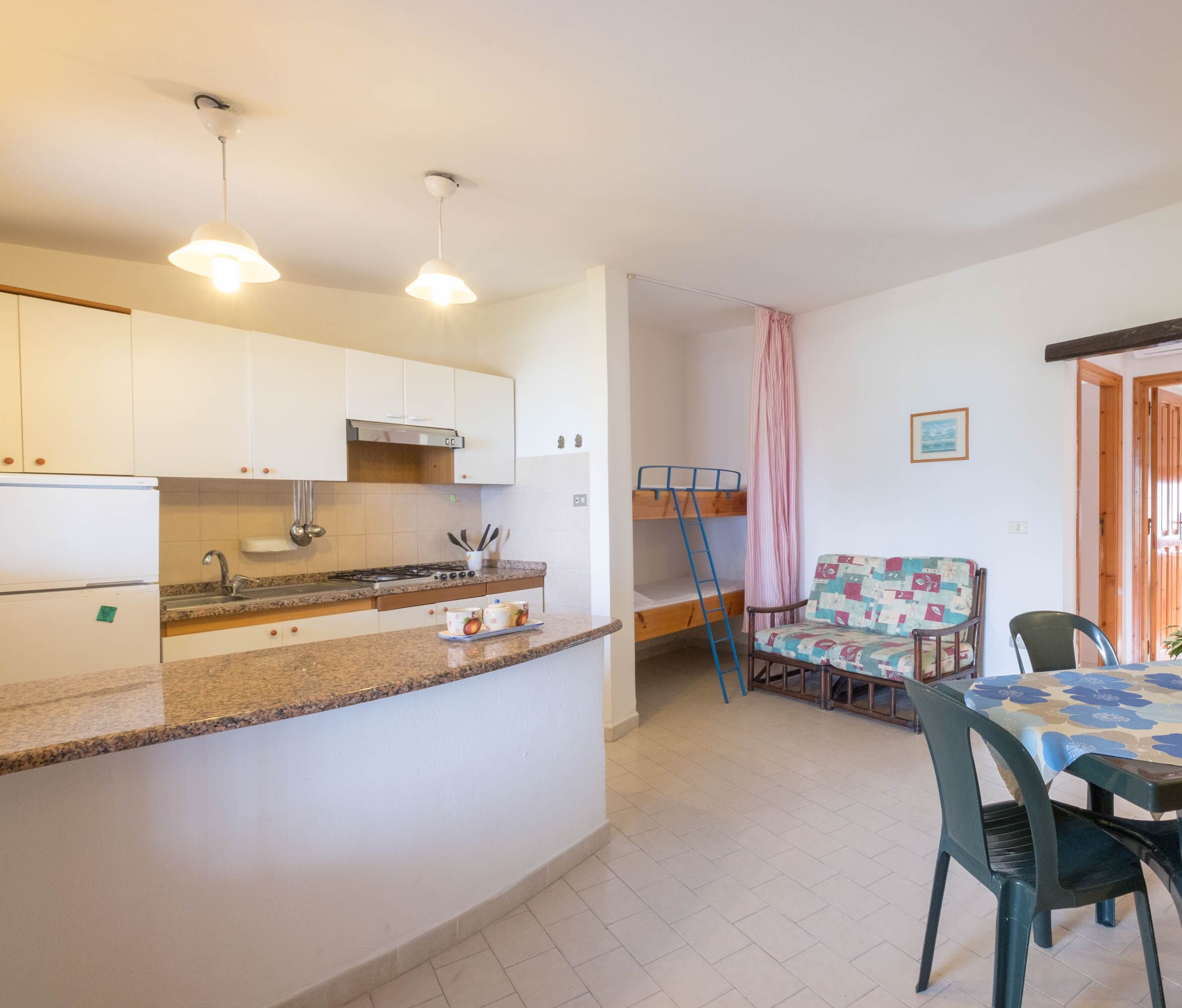 Una cocina compacta con armarios blancos, encimera de granito y estufa integrada. En la habitación contigua hay una cama litera con cortina rosa y una pequeña mesa con sillas.