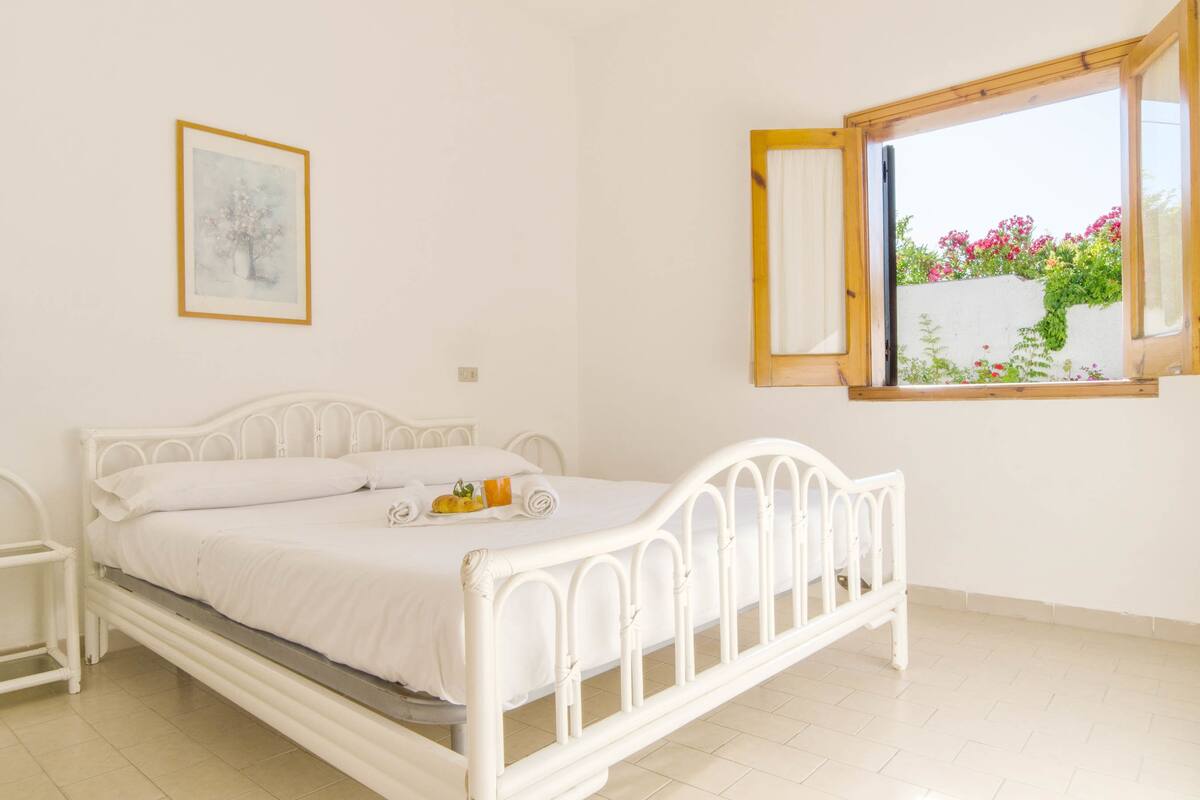 Una cama de metal blanca con tablero de cabecera de madera, una mesita de noche y una ventana con vista a flores.
