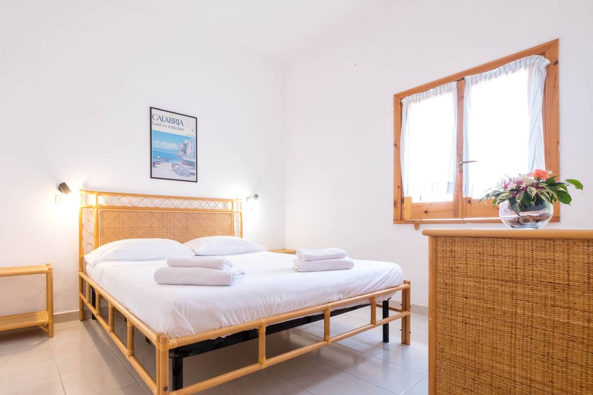Un dormitorio con cama, una mesita de noche pequeña, una mesita auxiliar de mimbre, una ventana con cortinas y un cuadro enmarcado en la pared.