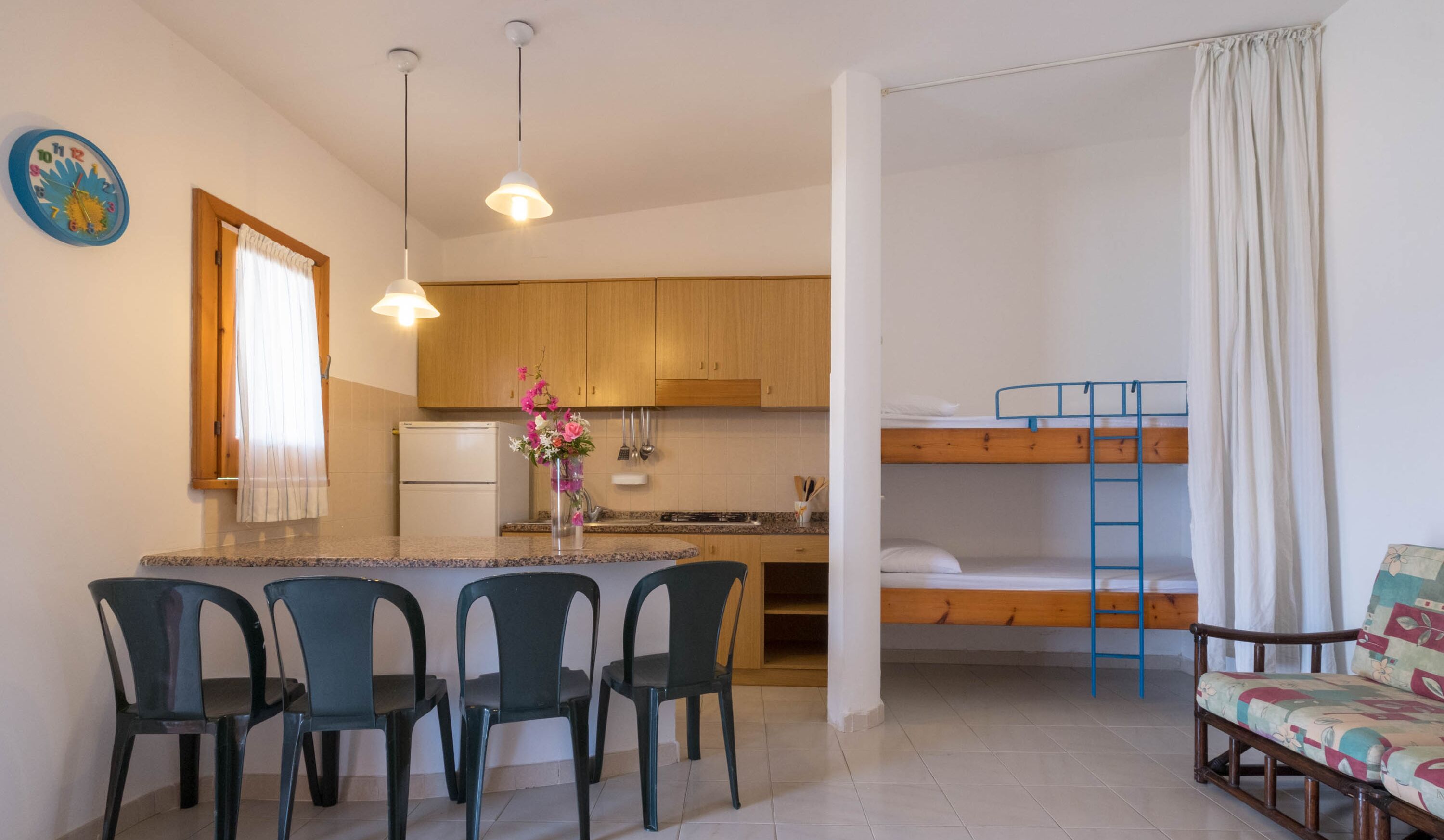 A compact kitchen with wooden cabinets, a refrigerator, and a dining area with four chairs. A bunk bed with a ladder is visible in the background.