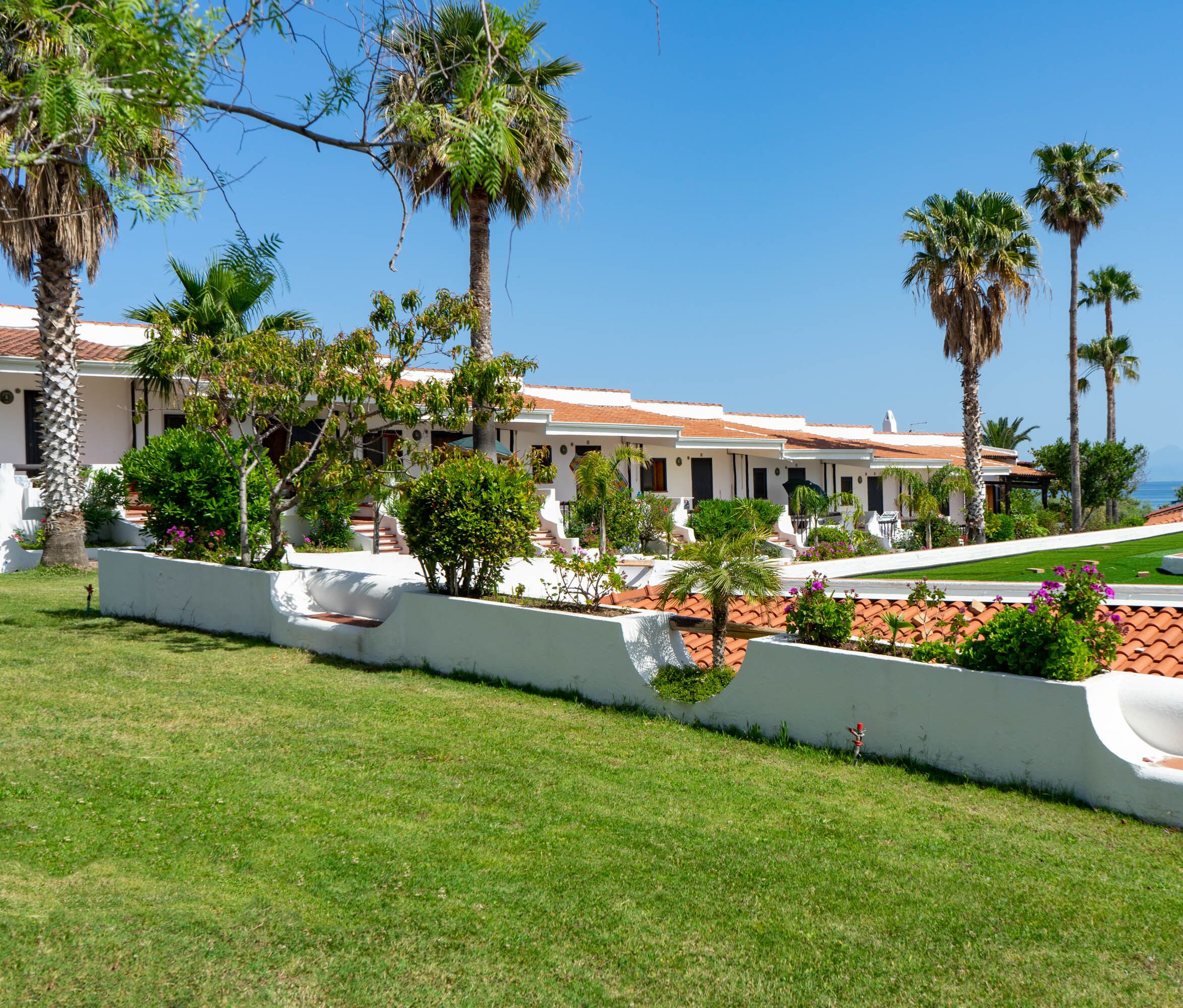 Un hotel con facciata bianca, tetto in terracotta e palme. In primo piano si vede una piscina e il cielo è di un azzurro intenso.