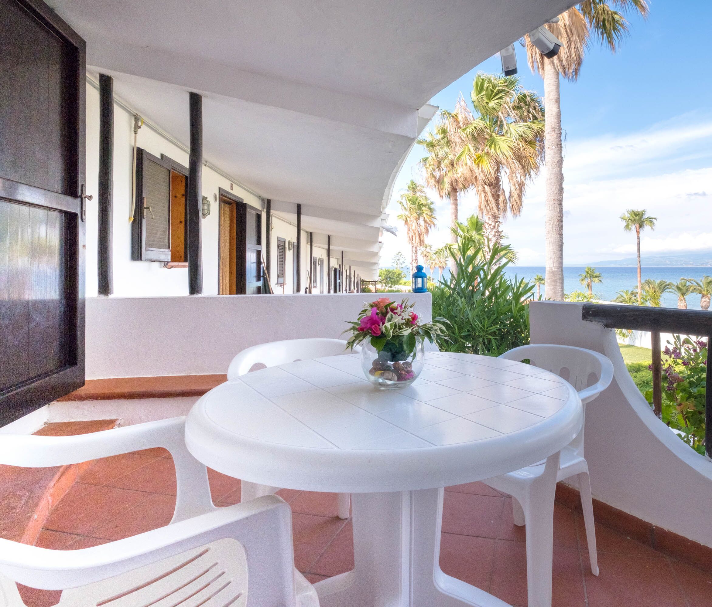 Ein Balkon mit rundem Tisch und Stühlen, einer Blumenvase und Blick auf das Meer und Palmen.