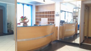 Una zona de recepción con un escritorio de madera curvo, un teléfono y un jarrón de flores. Se observan estanterías al fondo.