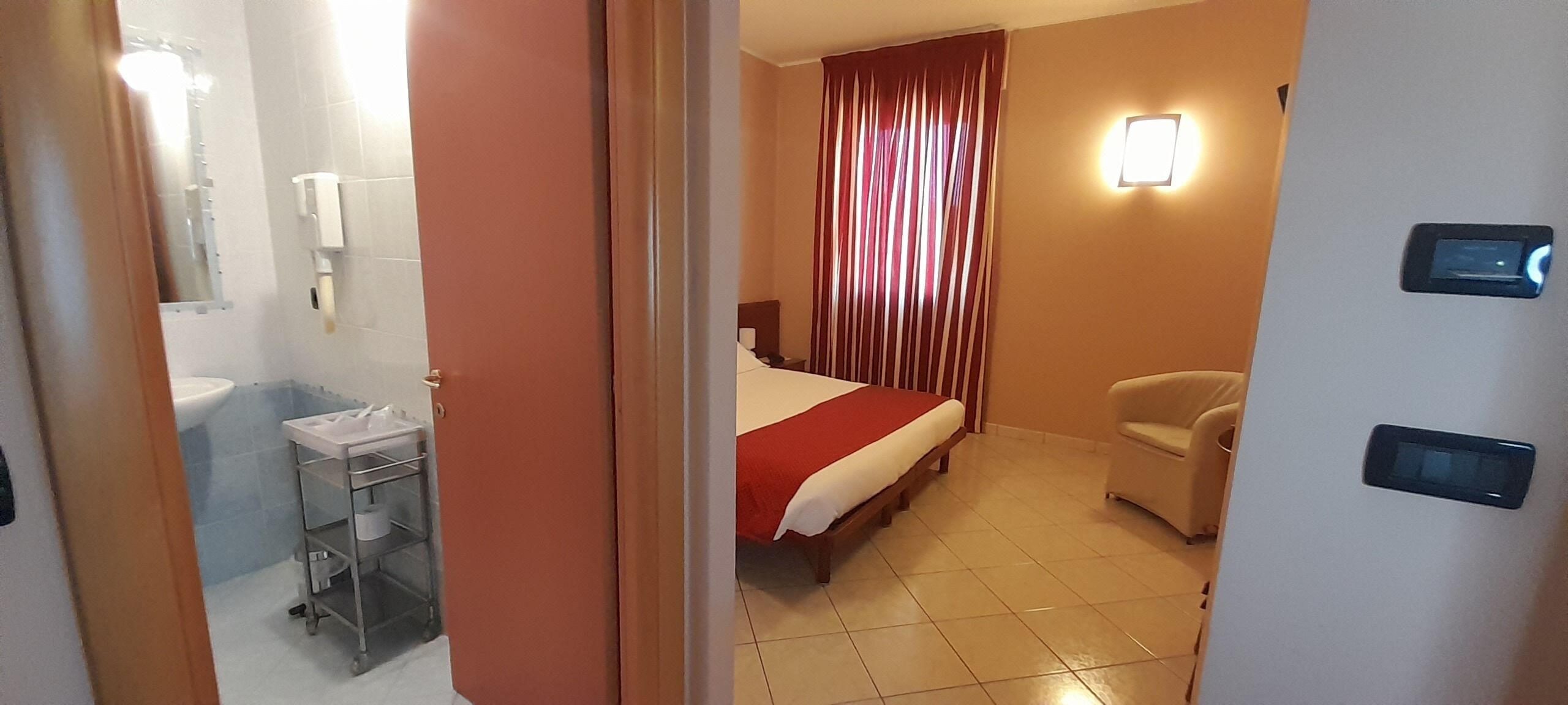 A hotel room with a bed, a chair, a bedside table, a nightstand, a wall-mounted light, a window with curtains, and a bathroom with a sink and a toilet.