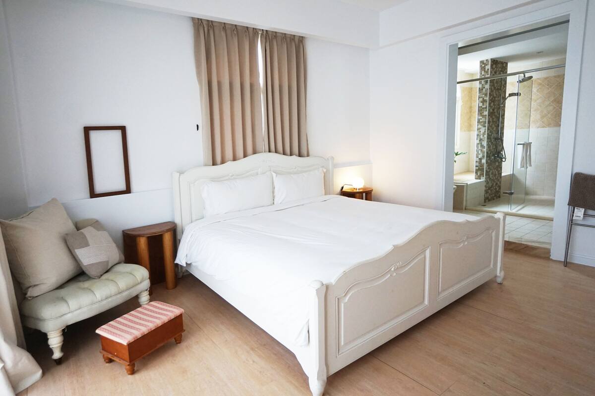 A bedroom with a large bed, a bedside table, a nightstand, a bench, a sofa, and a glass shower enclosure.