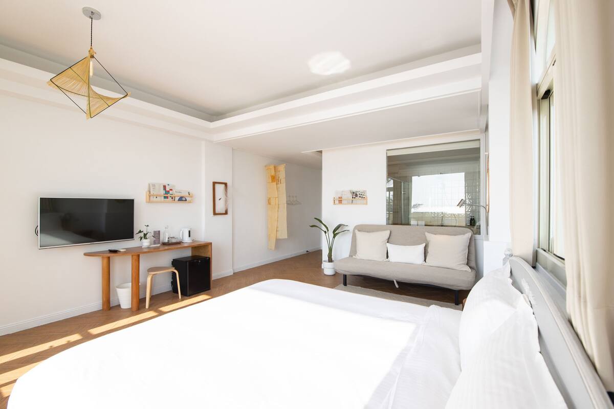 A modern hotel room with a large bed, a desk with a flat-screen TV, a sofa, and a large window.
