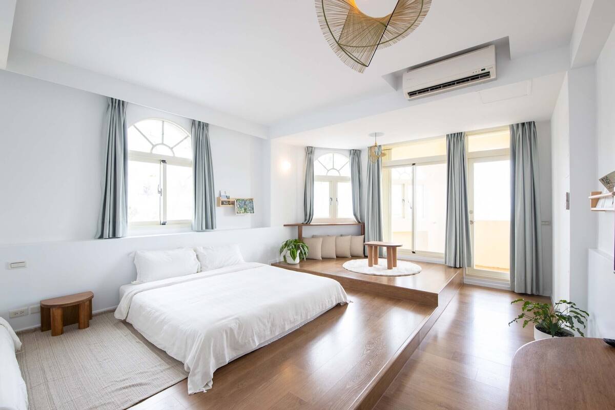 A modern bedroom with a large bed, a seating area with a round table, and a view of the outdoors through large windows.