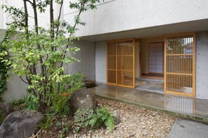 A traditional Japanese building with a concrete exterior, a wooden sliding door, and a small garden with rocks and plants.
