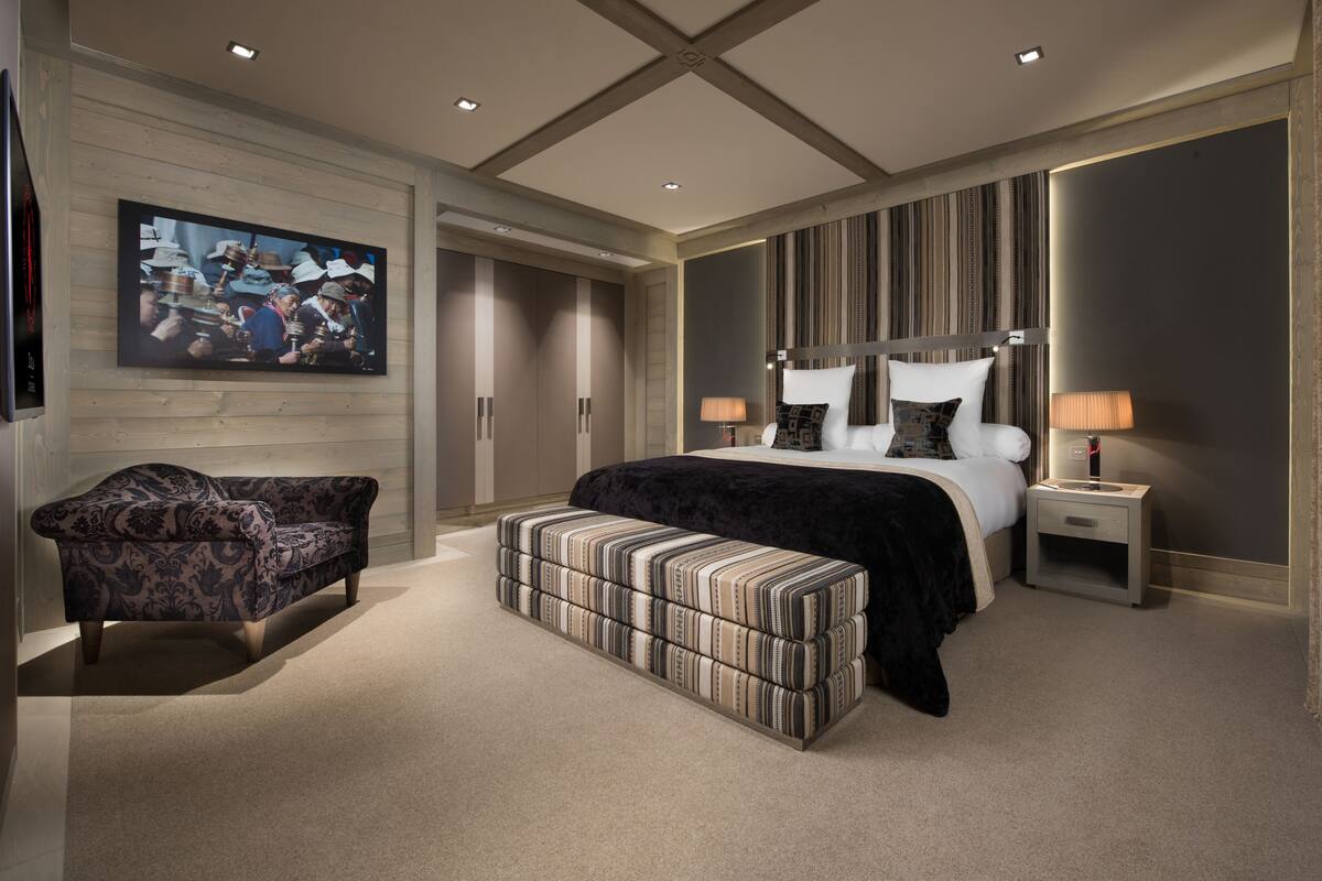 Quarto moderno com uma cama grande, um sofá, um banquinho, uma TV e uma mesa de cabeceira com luminárias.