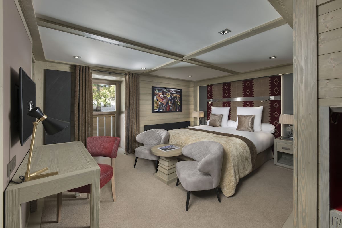 Quarto de hotel com uma cama, duas poltronas, uma mesa de trabalho, uma televisão e um quadro na parede.