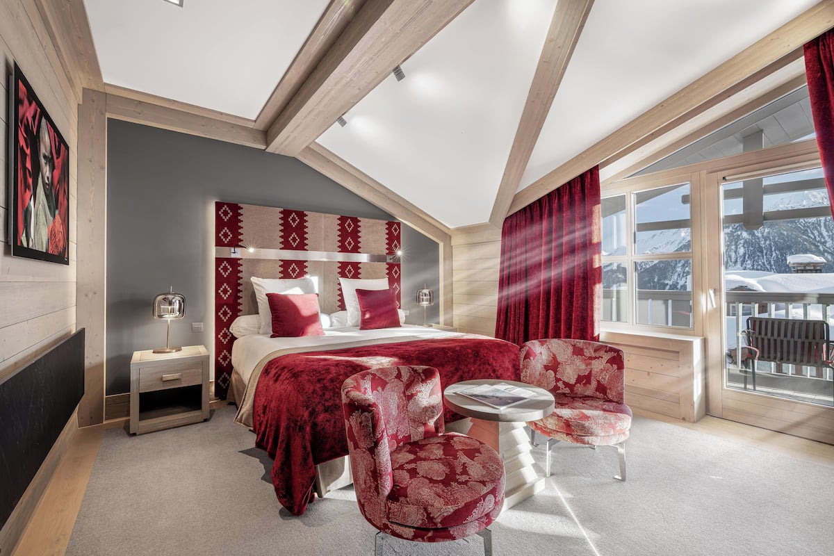 Quarto com uma cama grande, cabeceira estampada em vermelho e branco, mesa de cabeceira, poltrona estofada em vermelho, pequena mesa redonda e janela com vista para montanhas cobertas de neve.
