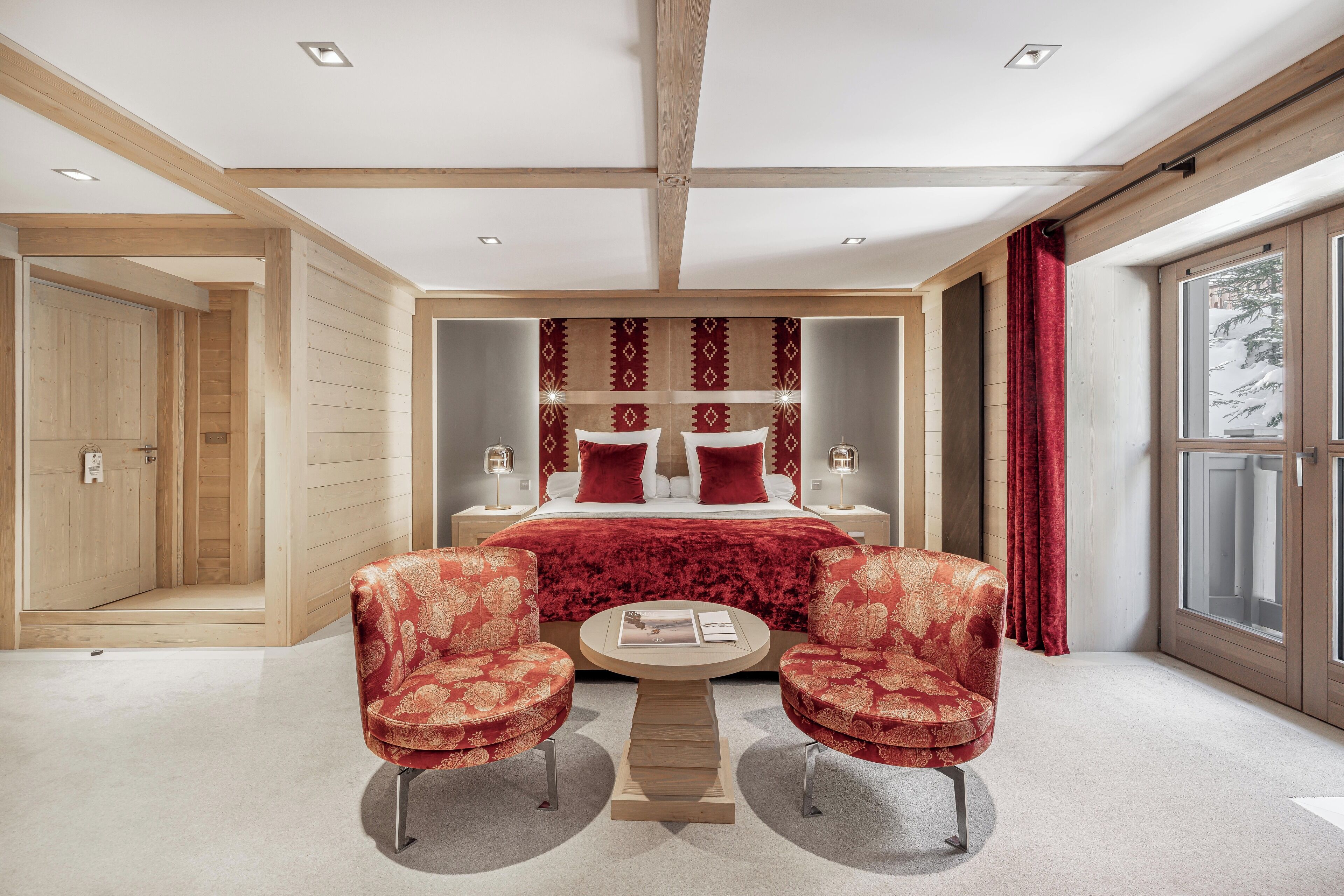 Une chambre à coucher moderne avec un grand lit, deux fauteuils rouges à revêtement en tissu, une table ronde en bois et une grande fenêtre avec des rideaux.
